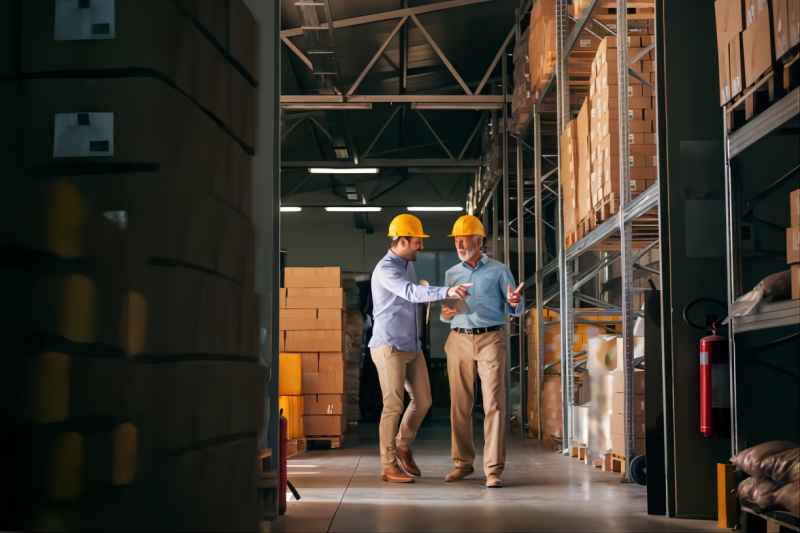 Workers walking in storage and pointing at boxes. In hands folder and tablet, on heads helmets. Workers walking in storage and pointing at bo(1)